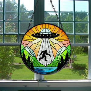 Puede incluir: Una ventana de vitral colgada de una cadena, que representa a un Bigfoot corriendo hacia un platillo volante en un paisaje montañoso colorido.
