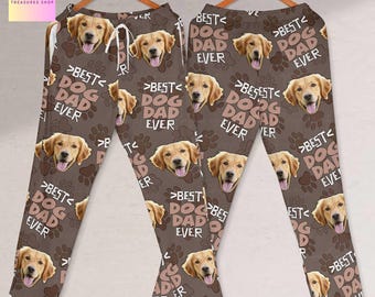 Może przedstawiać: Brązowe spodnie od piżamy z powtarzającym się wzorem twarzy golden retrievera, śladów łap i napisem "BEST DOG DAD EVER". Spodnie mają talię ze sznurkiem i są prezentowane na drewnianym wieszaku.