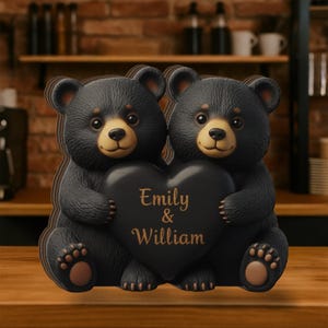 Pode incluir: Duas figuras de urso castanho-escuro sentadas lado a lado, segurando um grande coração preto entre elas. O coração apresenta o texto dourado Emily & William. Os ursos têm focinhos, orelhas internas e almofadas das patas castanho-claras, com pelo texturizado.