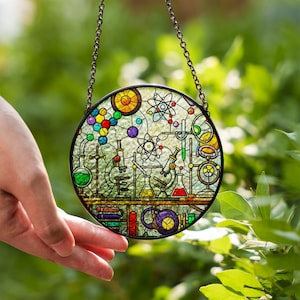 May include: A round, stained glass window featuring a science lab design with beakers, atoms, and a microscope. The window is framed in black and hangs from a chain. The design incorporates various colors, including red, yellow, green, and blue.