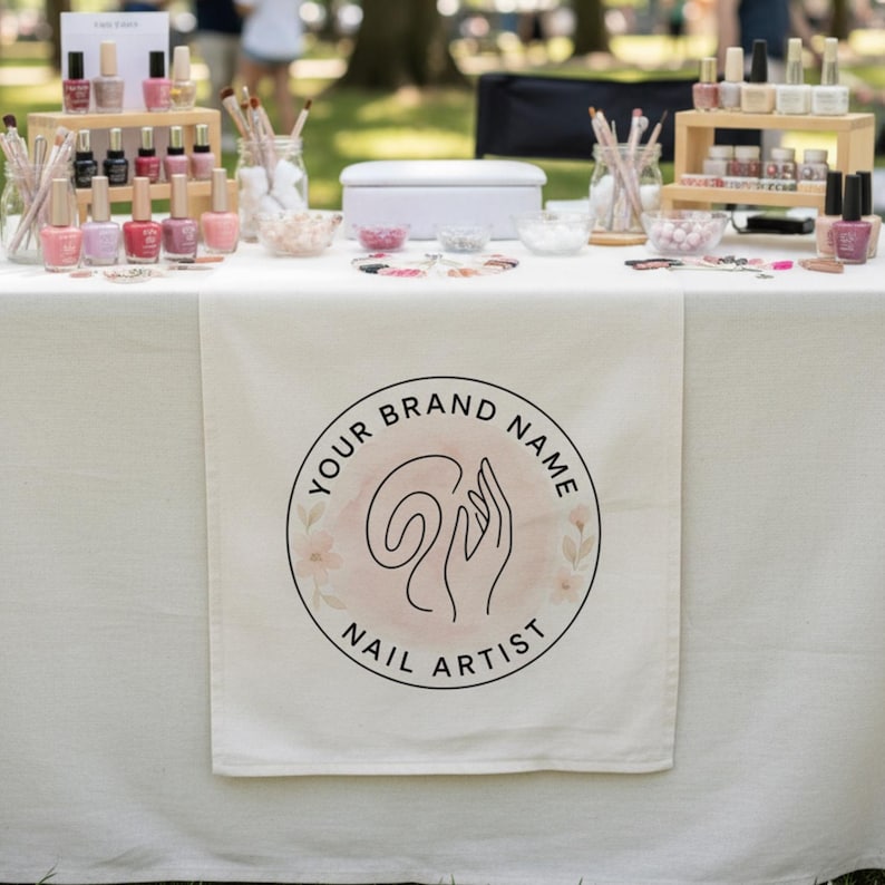 Puede incluir: Un camino de mesa de tela blanca con un logotipo circular que incluye el texto YOUR BRAND NAME NAIL ARTIST y una ilustraci&oacute;n lineal de una mano. La mesa est&aacute; dispuesta con varios esmaltes de u&ntilde;as, pinceles en frascos y cuencos con cuentas.