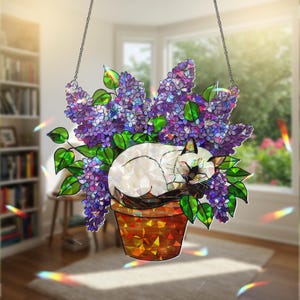May include: A stained glass window hanging features a white cat sleeping in a pot of purple flowers. The cat is surrounded by purple and green leaves. The window hanging is made of iridescent glass.
