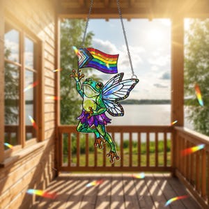 Può includere: Un acchiappasole in vetro colorato raffigurante una rana verde con ali di fata e una gonna viola, che tiene in mano una bandiera arcobaleno del Pride. L'acchiappasole è appeso a una catena d'argento. Lo sfondo mostra un portico in legno e un lago.