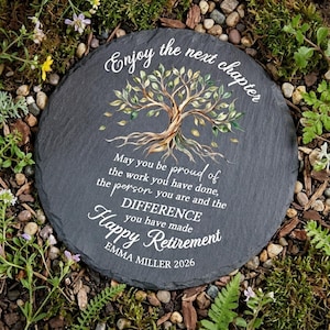Può includere: Una pietra per aiuole rotonda, grigio scuro, con le parole "Enjoy the next chapter" e "Happy Retirement" in corsivo bianco. Un'illustrazione di un albero è al centro. Il testo recita "May you be proud of the work you have done, the person you are and the DIFFERENCE you have made. EMMA MILLER 2026."