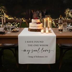 May include: A white table runner with the text "I HAVE FOUND THE ONE WHOM my soul loves - Song of Solomon 3:4". A two-tiered cake with berries sits on a wooden base. Lit candles and glasses are on the table, with fairy lights in the background.