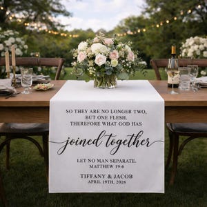 May include: White table runner with black script that reads "joined together" and wedding details. A floral arrangement of pink and white flowers sits in a glass vase. The table is set for a wedding reception.