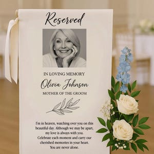 May include: A memorial sign with the word "Reserved" at the top. It features a black and white photo of a woman, with the text "IN LOVING MEMORY Olivia Johnson MOTHER OF THE GROOM". The sign includes a floral arrangement and a heartfelt message.