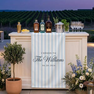 Puede incluir: Un camino de mesa a rayas azul claro y blanco sobre una barra blanca, con el texto impreso 'CHEERS TO The Williams 06.20.2026'. La barra contiene tres botellas de licor de vidrio, una cubitera de metal con hierbas y varias copas de cristal.