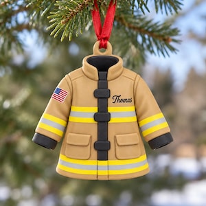 Può includere: Un ornamento a forma di giacca da vigile del fuoco color sabbia con chiusura nera, strisce riflettenti gialle e argento e due tasche frontali. Sulla manica destra c'è una piccola bandiera americana e il nome Thomas è scritto in nero.