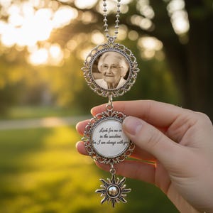 Op de afbeelding: Zilveren gedenkhanger met een foto van een vrouw in een sierlijk frame, hangend aan een ketting. De onderste hanger heeft de tekst "Look for me in the sunshine, for I am always with you." Een zonnesieraad hangt eronder.