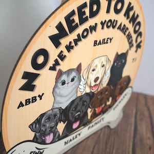 May include: A round wooden hanging sign with a jute rope, featuring the text NO NEED TO KNOCK WE KNOW YOU ARE HERE. The sign displays illustrations of a grey cat and several dogs, with the names ABBY, BAILEY, FINN, HALEY, and PAISLEY printed.