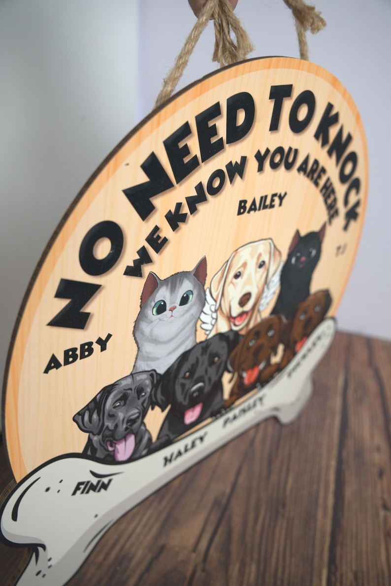 May include: A round wooden hanging sign with a jute rope, featuring the text NO NEED TO KNOCK WE KNOW YOU ARE HERE. The sign displays illustrations of a grey cat and several dogs, with the names ABBY, BAILEY, FINN, HALEY, and PAISLEY printed.