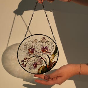 Puede incluir: Un adorno de ventana redondo de vidrio tintado que presenta un diseño floral de orquídeas blancas con acentos rosados y morados. El adorno está suspendido de una cadena negra.