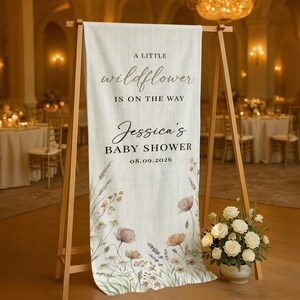 May include: A cream-colored baby shower sign on a wooden easel. The sign reads "A Little Wildflower Is On The Way" and "Jessica's Baby Shower 08.09.2026". Floral illustrations in muted tones decorate the bottom.