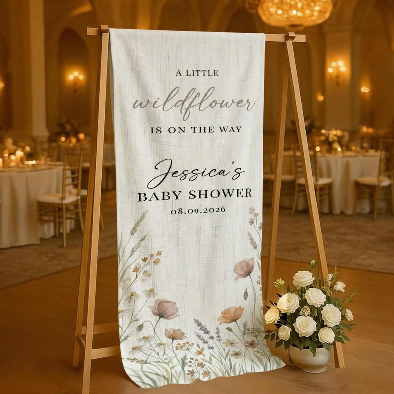 May include: A cream-colored baby shower sign on a wooden easel. The sign reads "A Little Wildflower Is On The Way" and "Jessica's Baby Shower 08.09.2026". Floral illustrations in muted tones decorate the bottom.