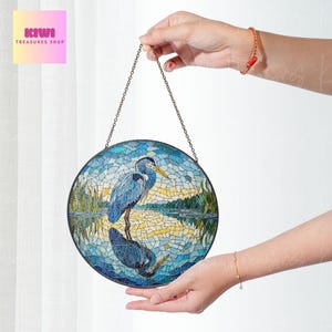 Könnte beinhalten: Ein rundes Buntglasfenster, das an einer Kette hängt. Das Fenster zeigt einen blauen Reiher, der in einem See steht, mit einer Spiegelung des Reihers im Wasser. Der Hintergrund ist eine Mosaik aus Blau, Grün und Gelb.