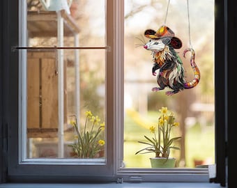 May include: A stained glass window hanging depicting a possum wearing a cowboy hat. The possum is colourful with shades of orange, yellow, red, and blue. The possum is hanging in front of a window with a view of a garden with yellow flowers.