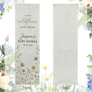 May include: A rectangular banner with the text "A little wildflower is on the way" and "Jessica's BABY SHOWER 08.09.2026". The banner has a floral design with wildflowers in shades of brown, yellow, and purple. The back of the banner is plain.