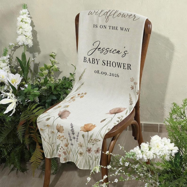 May include: A cream-colored baby shower banner draped over a wooden chair, featuring floral illustrations in muted tones. The banner reads "wildflower is on the way" and "Jessica's BABY SHOWER 08.09.2026". Decorative flowers and greenery surround the chair.