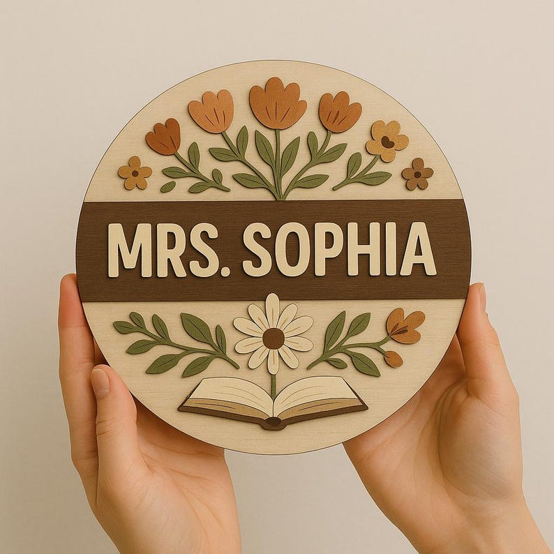 May include: A round wooden sign with the text "MRS. SOPHIA" in a dark brown band. The sign is decorated with flowers, leaves, and an open book. The sign is held by two hands.