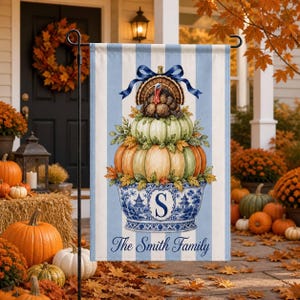 May include: A vertical garden flag with light blue and white stripes featuring a blue and white porcelain bowl filled with stacked pumpkins, autumn leaves, and a turkey topped with a blue bow. The bowl displays the letter S and the text The Smith Family.