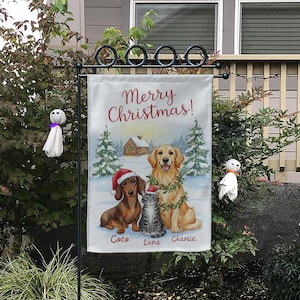 May include: A festive garden flag with a watercolour illustration of a dachshund, a cat, and a golden retriever in a snowy scene. The flag reads "Merry Christmas!" in red script, with the pets' names below.