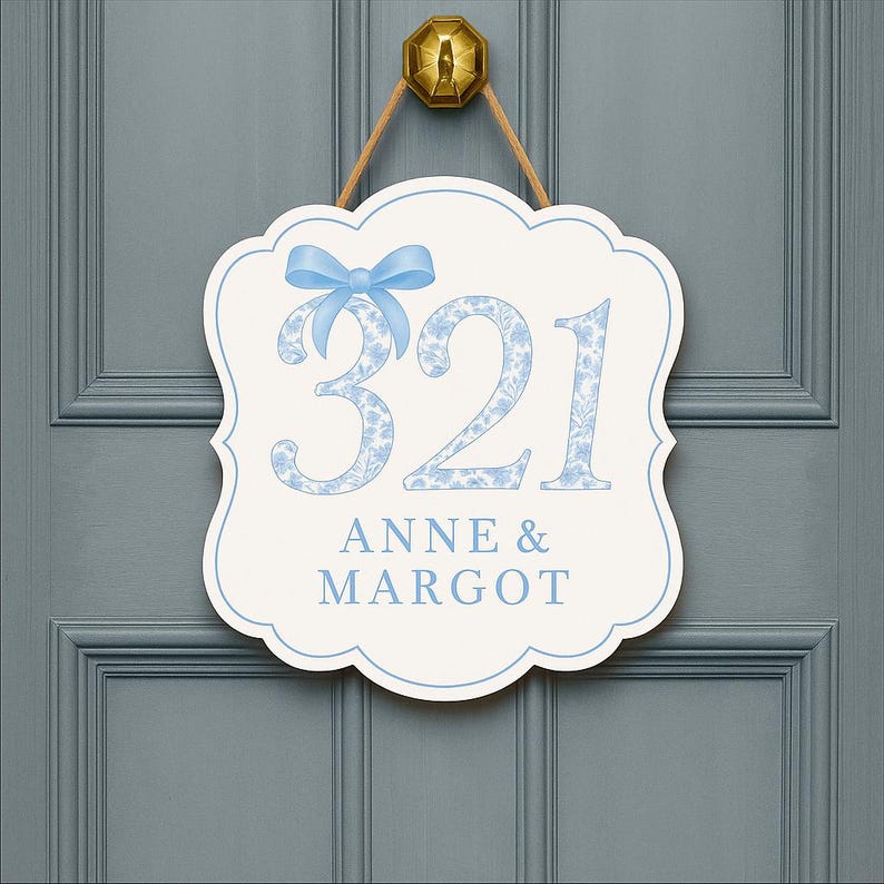 May include: A white, scalloped-edge wall sign featuring the number 321 in a blue floral pattern, topped with a light blue bow. Below the number, the text ANNE & MARGOT is printed in blue. The sign hangs from a gold-colored hook by a tan cord.