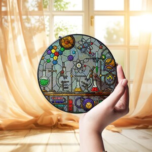 May include: A round stained glass panel featuring a science lab design with beakers, microscopes, and atom models. The panel has a black border and is held in front of a window with sunlight streaming through.