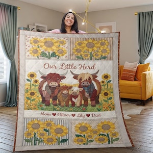 May include: A quilted blanket featuring a sunflower and cow design. The blanket has the text "Our Little Herd" and names below the cow family. The blanket is cream-colored with a brown border.