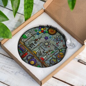 May include: A round, stained glass ornament featuring a science lab design with beakers, test tubes, and atom models in various colors. The ornament is in a cardboard box with white padding, ready for gifting.