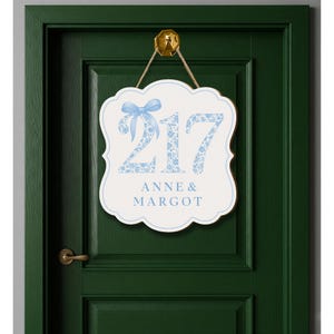 May include: A white scalloped wall sign featuring the number 217 in a light blue floral pattern with a matching blue bow graphic above the numbers. Below the number, the text ANNE & MARGOT is printed in blue. The sign hangs from a gold hook.