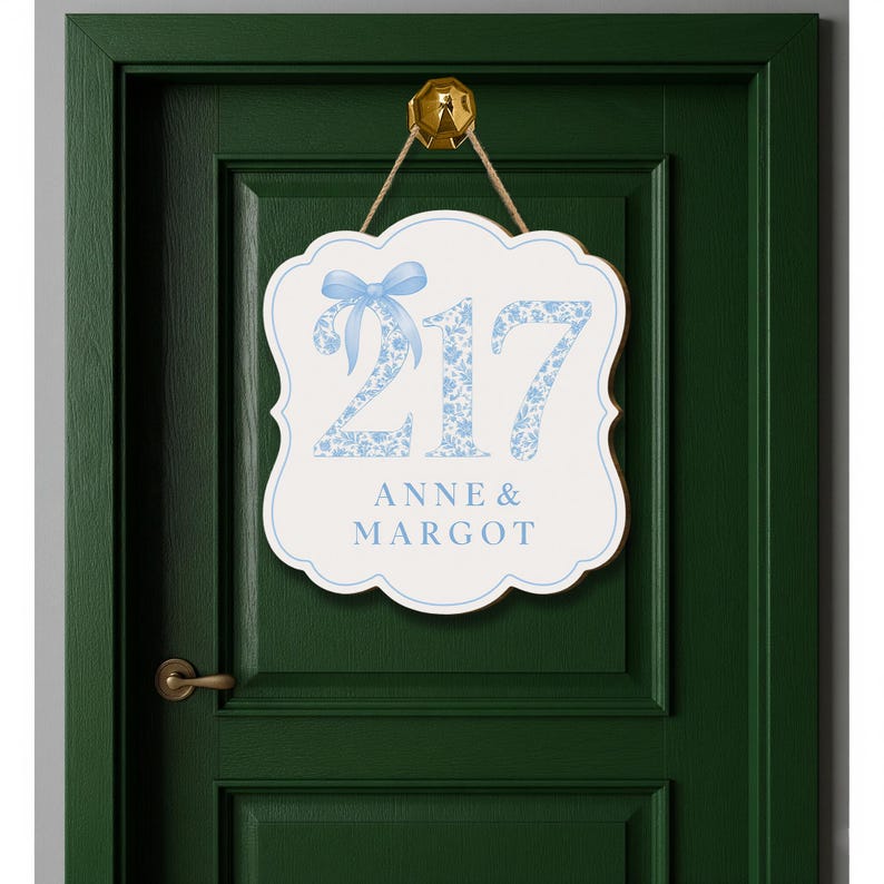 May include: A white scalloped wall sign featuring the number 217 in a light blue floral pattern with a matching blue bow graphic above the numbers. Below the number, the text ANNE & MARGOT is printed in blue. The sign hangs from a gold hook.