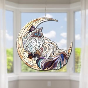May include: A stained glass cat suncatcher featuring a long-haired cat design, resting on a crescent moon. The cat has blue eyes and a multi-coloured coat. The moon is made of beige and brown glass, and hangs from a silver chain.