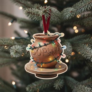 May include: A brown ceramic ornament shaped like a pottery vase, with the name "SOPHIA" carved into the front. The ornament is decorated with colorful Christmas lights and a red ribbon for hanging. The ornament is on a wooden base.