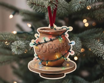 Può includere: Un ornamento in ceramica marrone a forma di vaso di terracotta, con il nome "SOPHIA" scolpito sul davanti. L'ornamento è decorato con luci natalizie colorate e un nastro rosso per appenderlo. L'ornamento è su una base di legno.