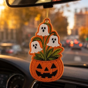 May include: A hanging car ornament featuring an embroidered orange jack-o'-lantern base with a black carved face. Three white ghost-shaped flowers with black eyes and mouths emerge from the pumpkin, surrounded by green leaves, suspended by a gold cord.