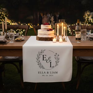 May include: A white table runner with a black floral wreath design, the initials "E & L", and the names "Ella & Liam" with the date "09. 14. 2025". A two-tiered cake with berries sits on a wooden base. Candles and glasses are on the wooden table.