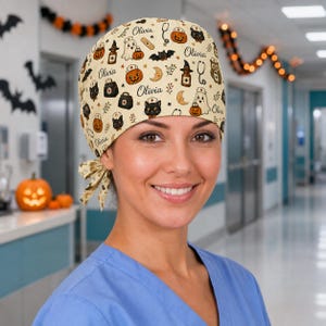 May include: A cream-coloured scrub cap featuring a repeating Halloween-themed pattern of pumpkins, ghosts, black cats, bats, stethoscopes, and medical bags, with the name Olivia printed in black script throughout the fabric and a side tie.
