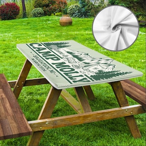 May include: A picnic table with a custom-printed cover featuring the text "Camp Molly" in green. The cover includes a mountain and tree design, with the location "Saugatuck, MI" and the date "March 2026". The table is made of wood and set on a grassy lawn.