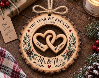 May include: A round wooden ornament with bark edges, featuring two interlocking hearts engraved with the names Olivia and Mason. The text reads THE YEAR WE BECAME 6.21.2026 and Mr. & Mrs. It is decorated with pine branches, red berries, and pinecones.