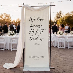 Puede incluir: Una pancarta de boda de color crema con texto negro que dice "We love because He first loved us" y "- 1 John 4:19". La pancarta tambi&eacute;n incluye los nombres "Kinsey & Sam" y la fecha "09.13.2026".