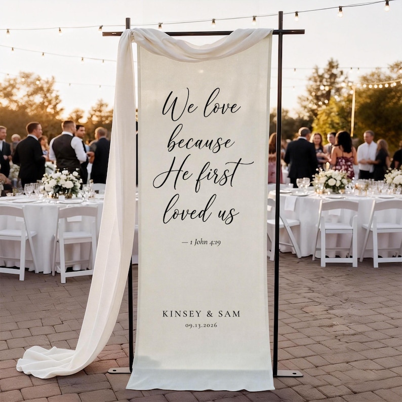 Puede incluir: Una pancarta de boda de color crema con texto negro que dice "We love because He first loved us" y "- 1 John 4:19". La pancarta tambi&eacute;n incluye los nombres "Kinsey & Sam" y la fecha "09.13.2026".