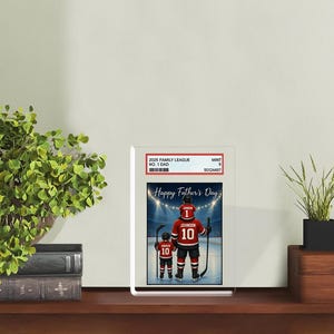May include: A framed print featuring a hockey-themed design for Father's Day. The image shows a father and child in hockey uniforms with the text "Happy Father's Day". The father's jersey reads "10 JOHNSON", and the child's reads "10 ANDREW".