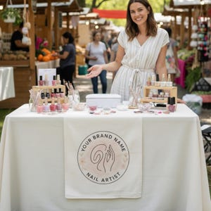May include: A nail artist's booth at an outdoor market, featuring a variety of nail polish colors displayed on wooden shelves. The table has a white tablecloth and a banner with the text "YOUR BRAND NAME NAIL ARTIST". A woman stands behind the table.