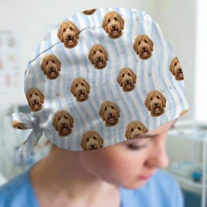 May include: A scrub cap featuring a repeating pattern of golden-brown doodle dog faces on a light blue and white wavy striped background, accented with small grey paw prints. The cap includes side ties for an adjustable fit.