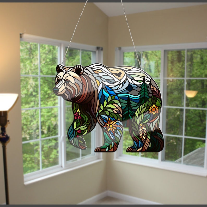 Puede incluir: Una silueta de oso de vidrio coloreado con un dise&ntilde;o colorido de una escena de bosque, que incluye &aacute;rboles, monta&ntilde;as y flores. El oso est&aacute; colgado de una cadena.