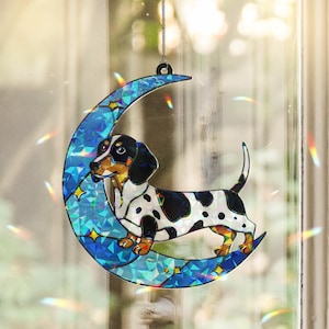 May include: A stained glass suncatcher featuring a black and white dachshund standing on a crescent moon. The moon is iridescent blue with silver stars and sparkles.