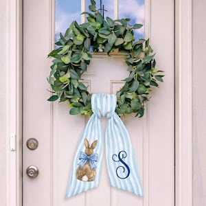 Op de afbeelding: Een groene eucalyptuskrans hangt aan een witte deur. Een blauw-wit gestreept lint is aan de krans bevestigd, met een bruin konijn met een blauwe strik en een cursieve letter "S".