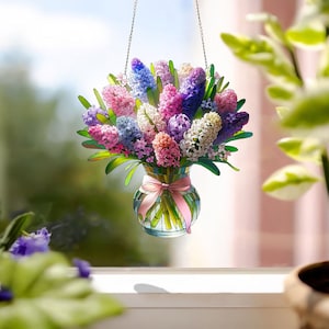 Puede incluir: Un ramo de jacintos rosas, morados y blancos en un jarrón de vidrio transparente con una cinta rosa. El jarrón está colgado de una cadena plateada.