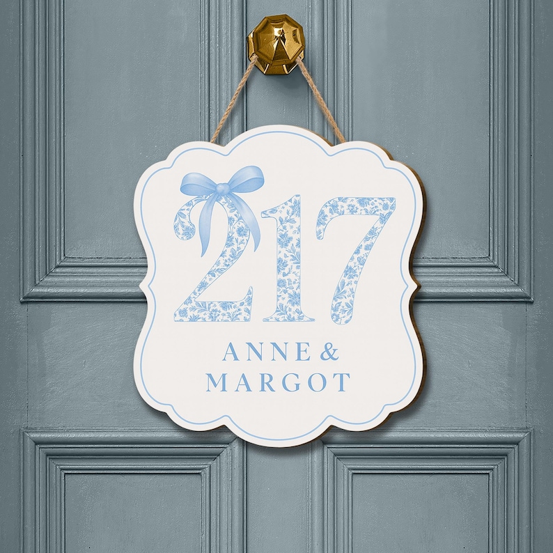May include: A white scalloped wall sign with a light blue border, featuring the number 217 in a blue floral pattern with a light blue bow on the number 2. Below the number, the text ANNE & MARGOT is printed in light blue capital letters.
